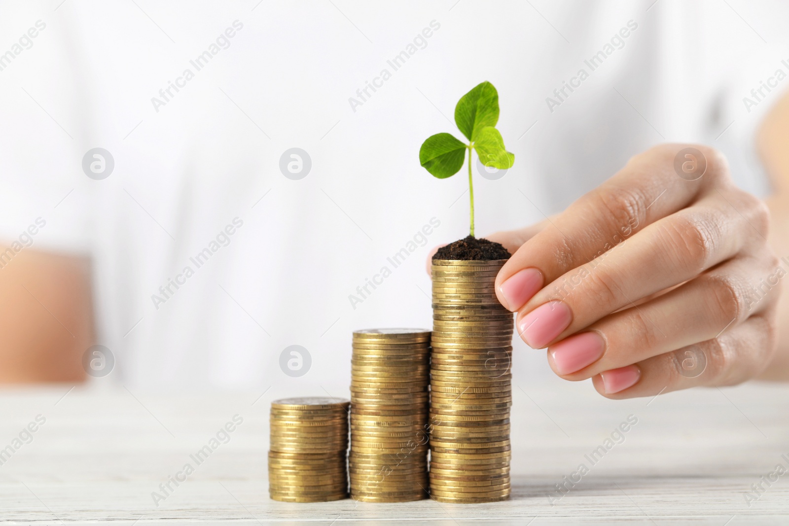 Photo of Woman putting coins with green sprout onto stack at white table, closeup. Investment concept