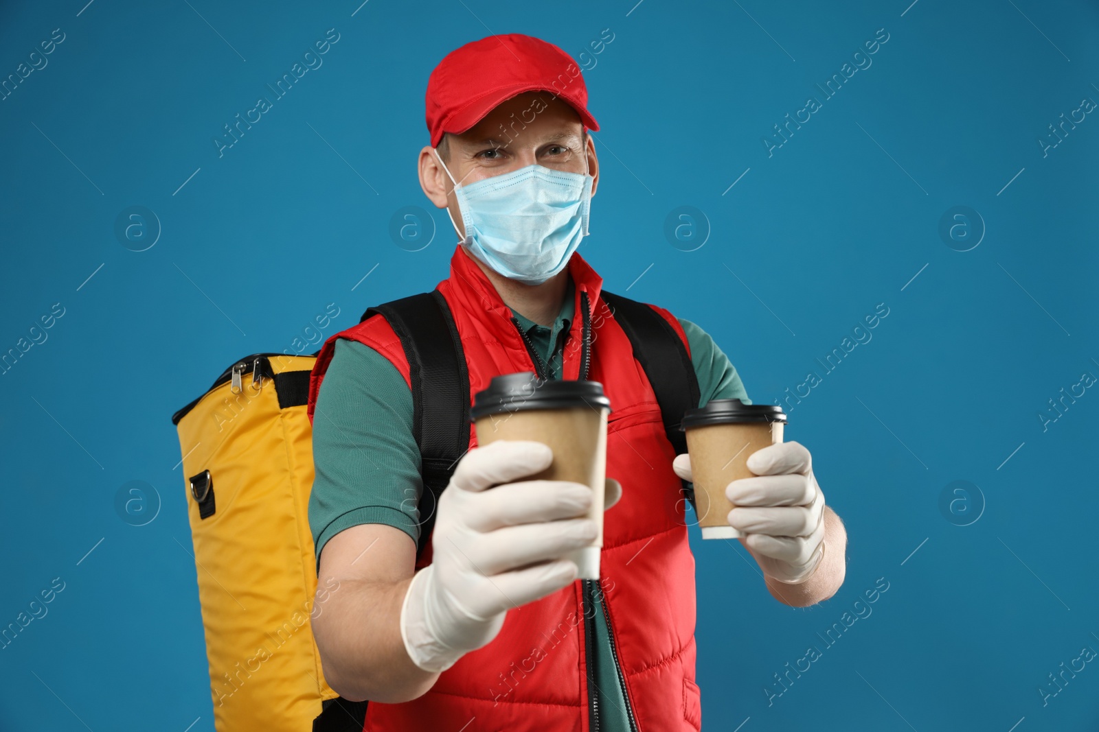 Photo of Courier in protective gloves and mask holding drinks on blue background. Delivery service during coronavirus quarantine