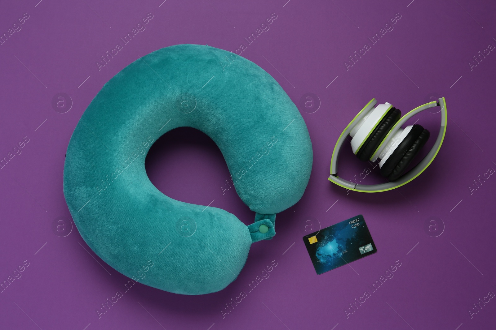 Photo of Turquoise travel pillow, headphones and credit card on purple background, flat lay