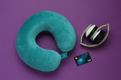 Photo of Turquoise travel pillow, headphones and credit card on purple background, flat lay