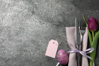 Cutlery set, painted egg and beautiful flower on grey table, flat lay with space for text. Easter celebration