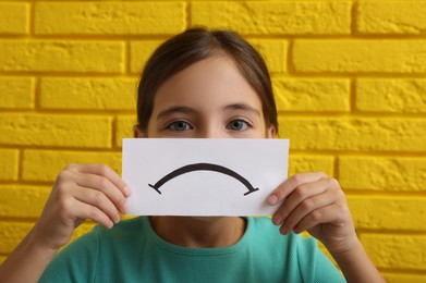 Sad face. Little girl showing feelings with card against yellow brick wall