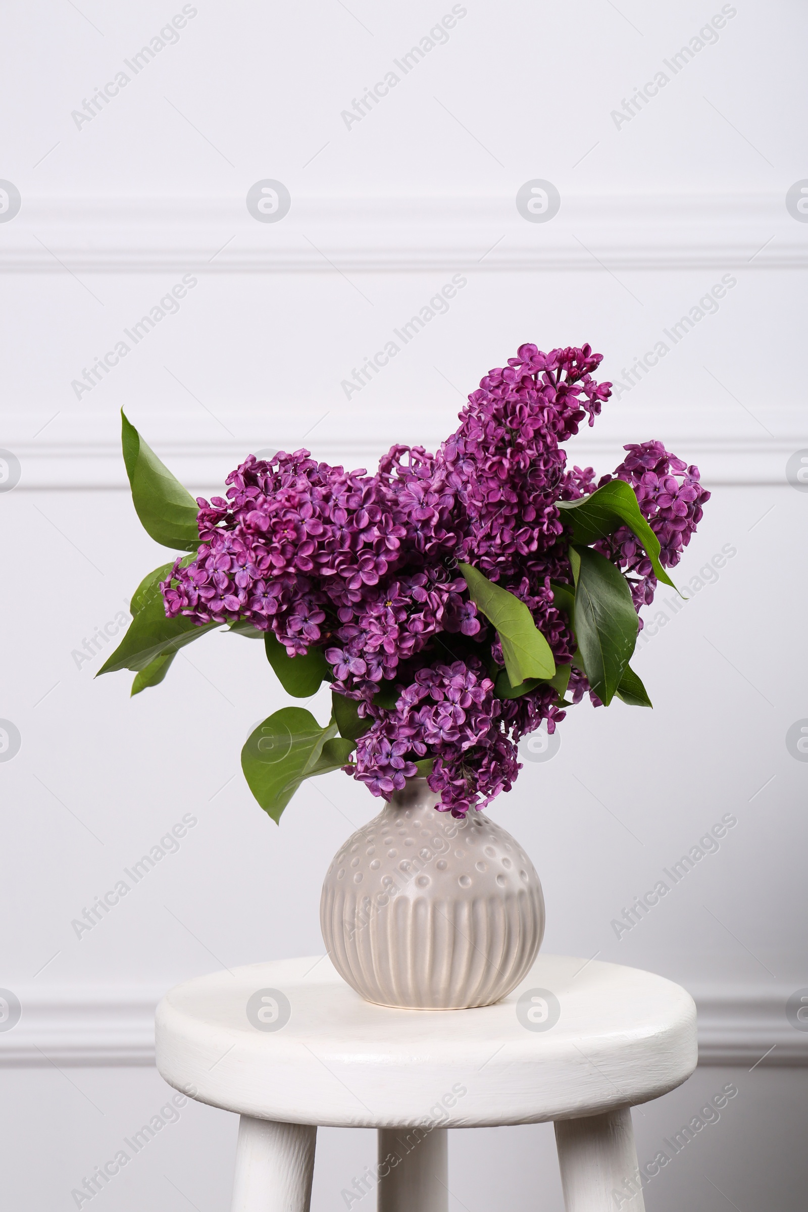 Photo of Beautiful lilac flowers in vase on white table near wall