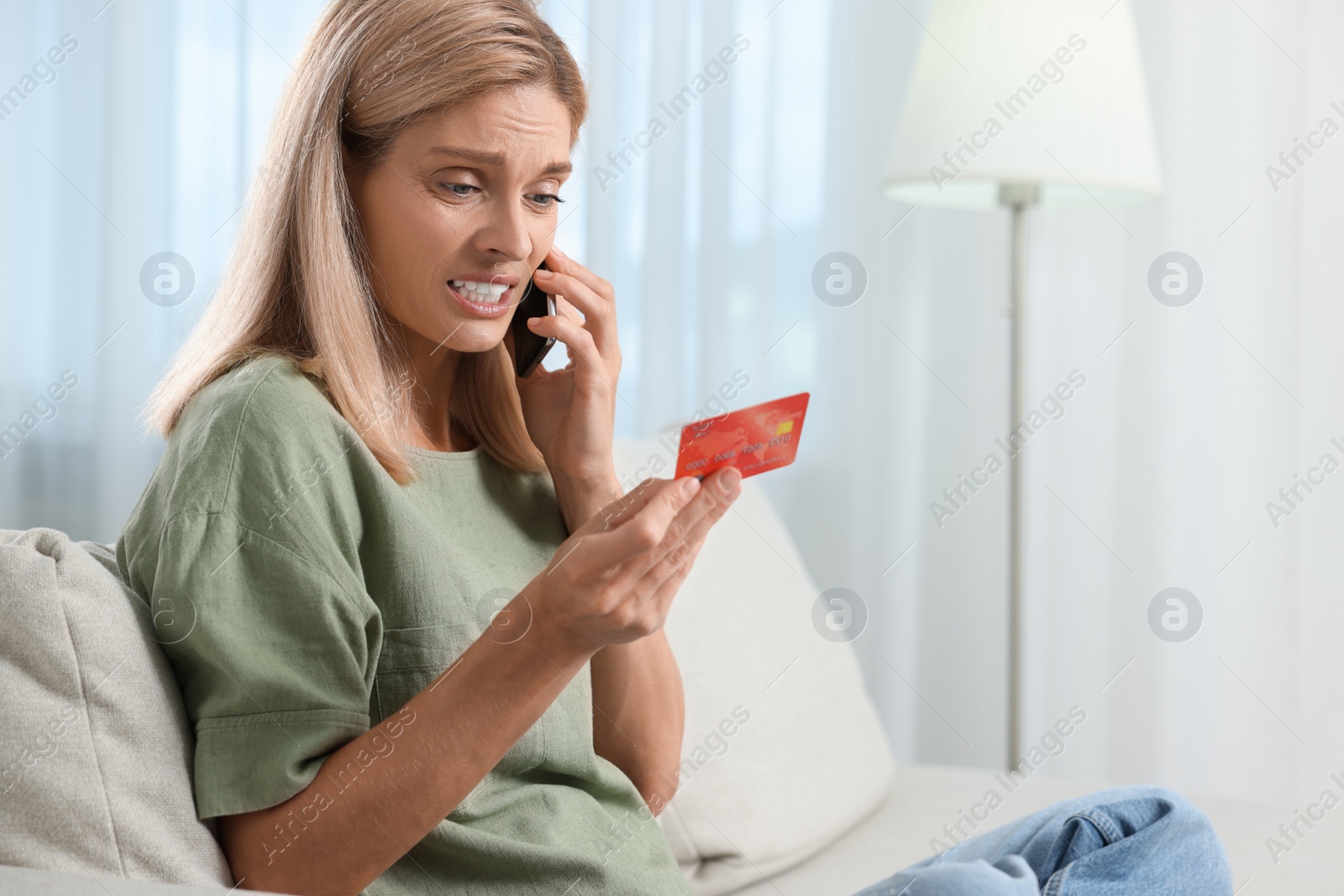 Photo of Emotional woman with credit card talking on smartphone at home, space for text. Be careful - fraud