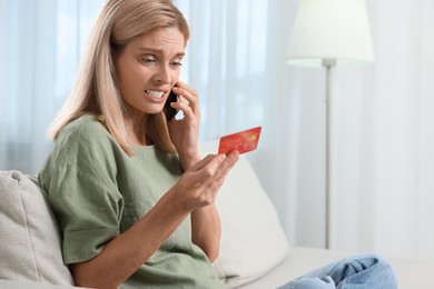 Emotional woman with credit card talking on smartphone at home, space for text. Be careful - fraud