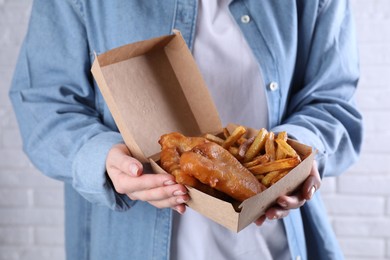 Photo of Woman holding fish and chips in paper box near white brick wall, closeup