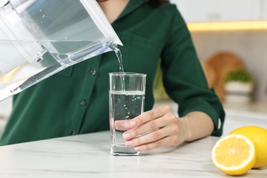 Photo of Woman pouring water from filter jug into glass in kitchen, closeup