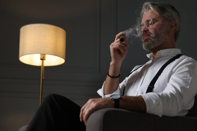 Photo of Bearded man smoking cigar in armchair indoors. Space for text