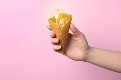 Photo of Woman holding waffle ice cream cone with light bulb on pink background, closeup