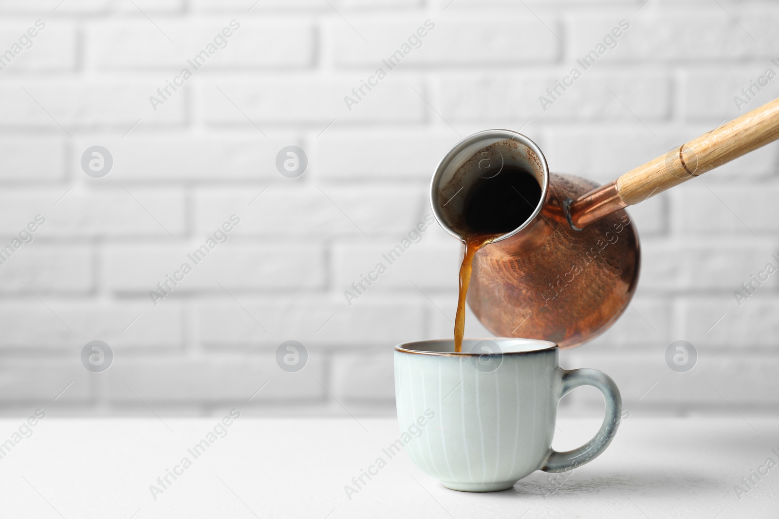 Photo of Turkish coffee. Pouring brewed beverage from cezve into cup at table against white brick wall. Space for text