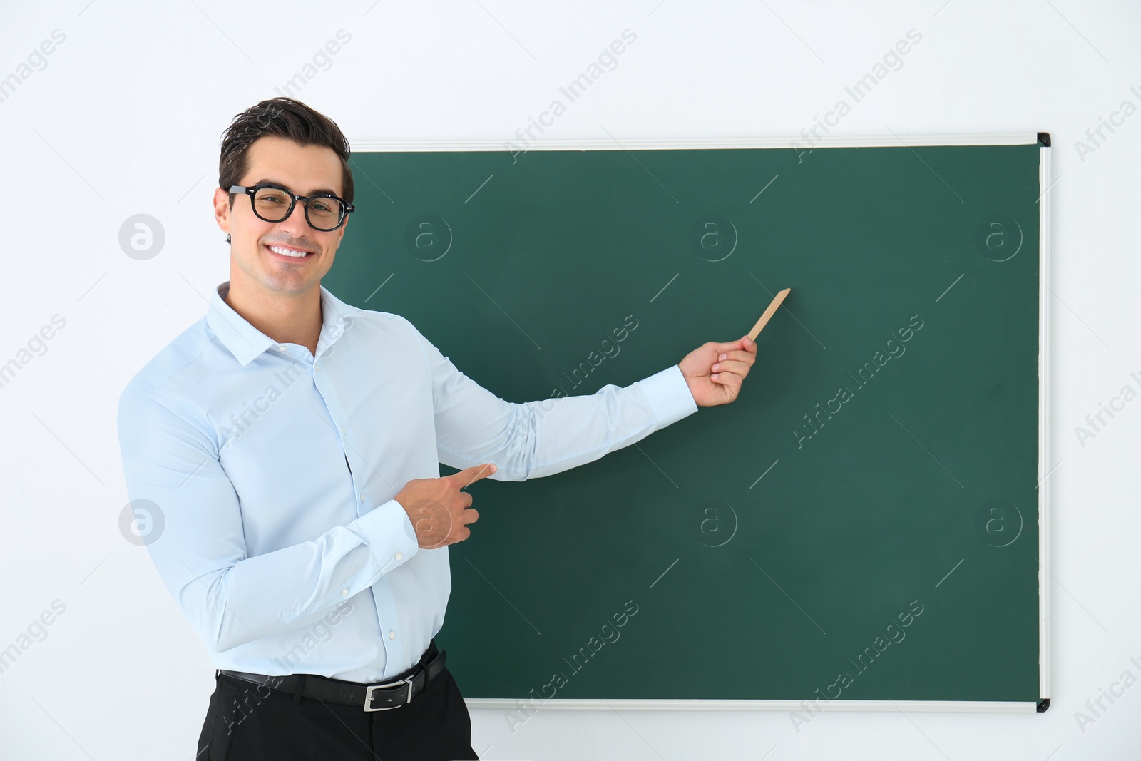 Photo of Young teacher with ruler near blank chalkboard in classroom. Space for text