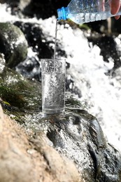 Woman pouring water from plastic bottle into glass near stream outdoors, closeup