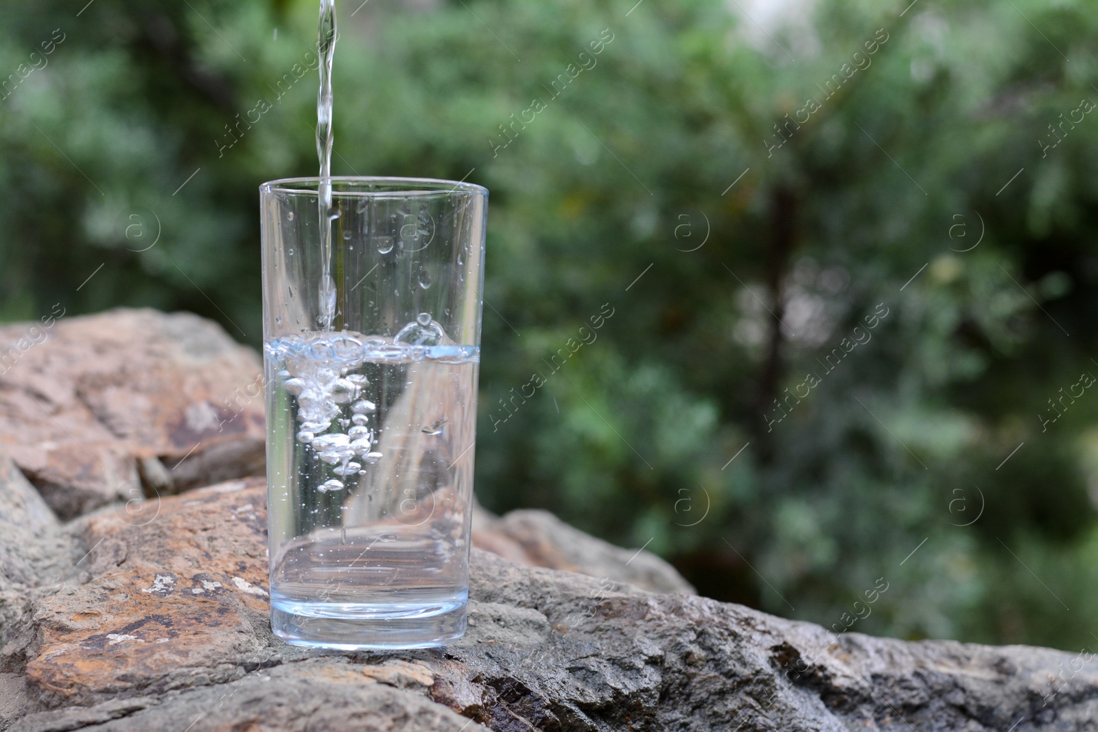 Photo of Pouring pure water into glass on stone outdoors, space for text