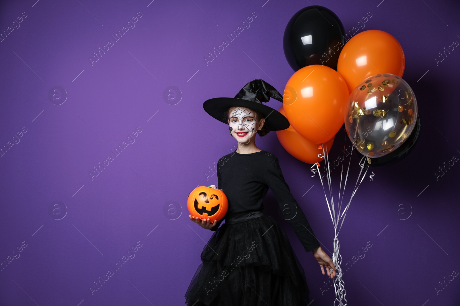 Photo of Cute little girl with pumpkin candy bucket and balloons wearing Halloween costume on purple background. Space for text