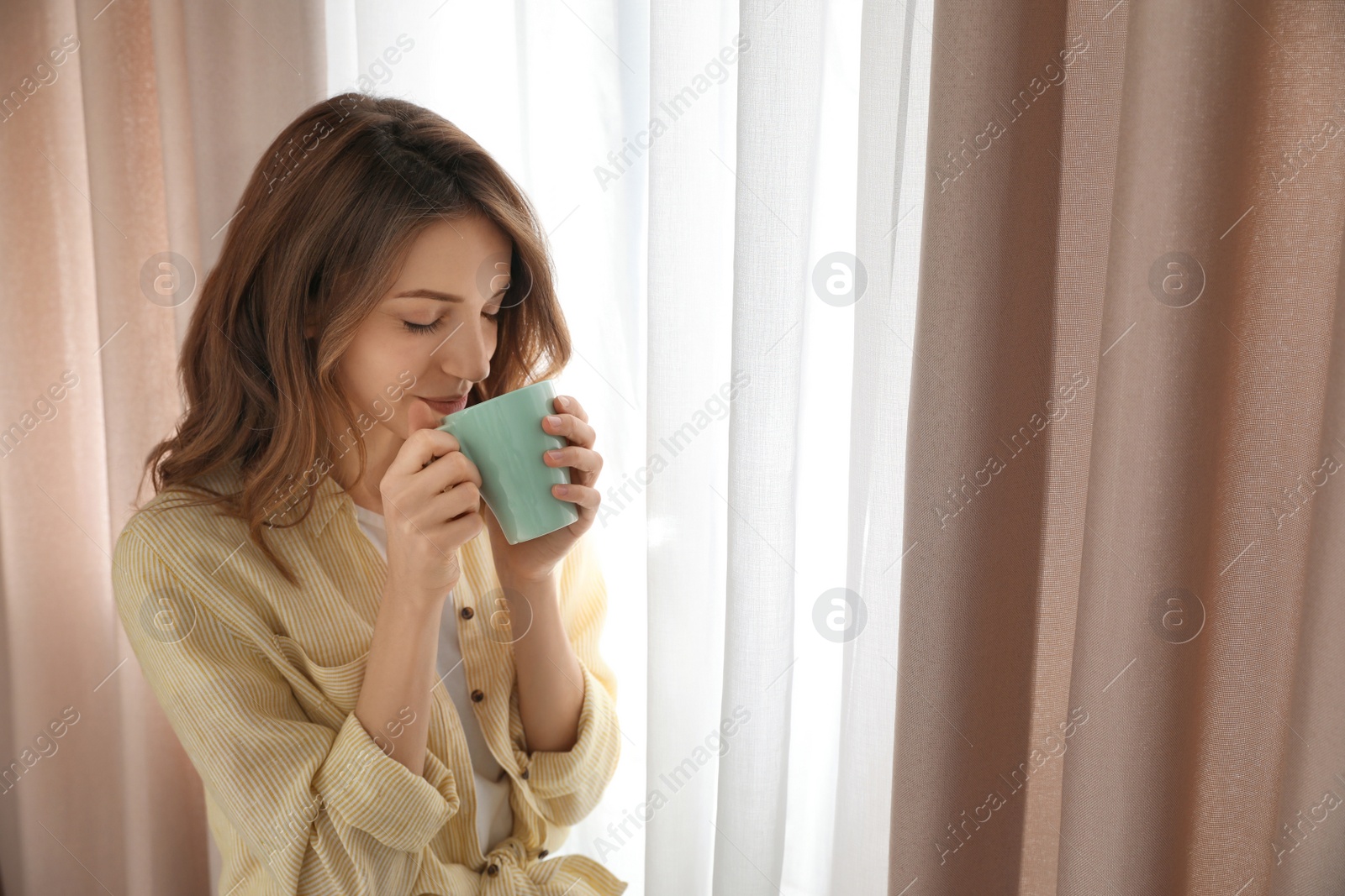 Photo of Woman drinking coffee near window with beautiful curtains at home