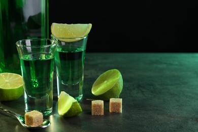 Absinthe in shot glasses, spoon, lime and brown sugar cubes on gray textured table against black background, space for text. Alcoholic drink