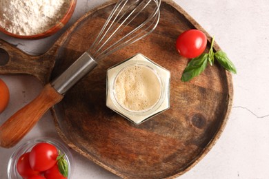 Photo of Pizza dough starter in glass jar, products and whisk on gray textured table, flat lay