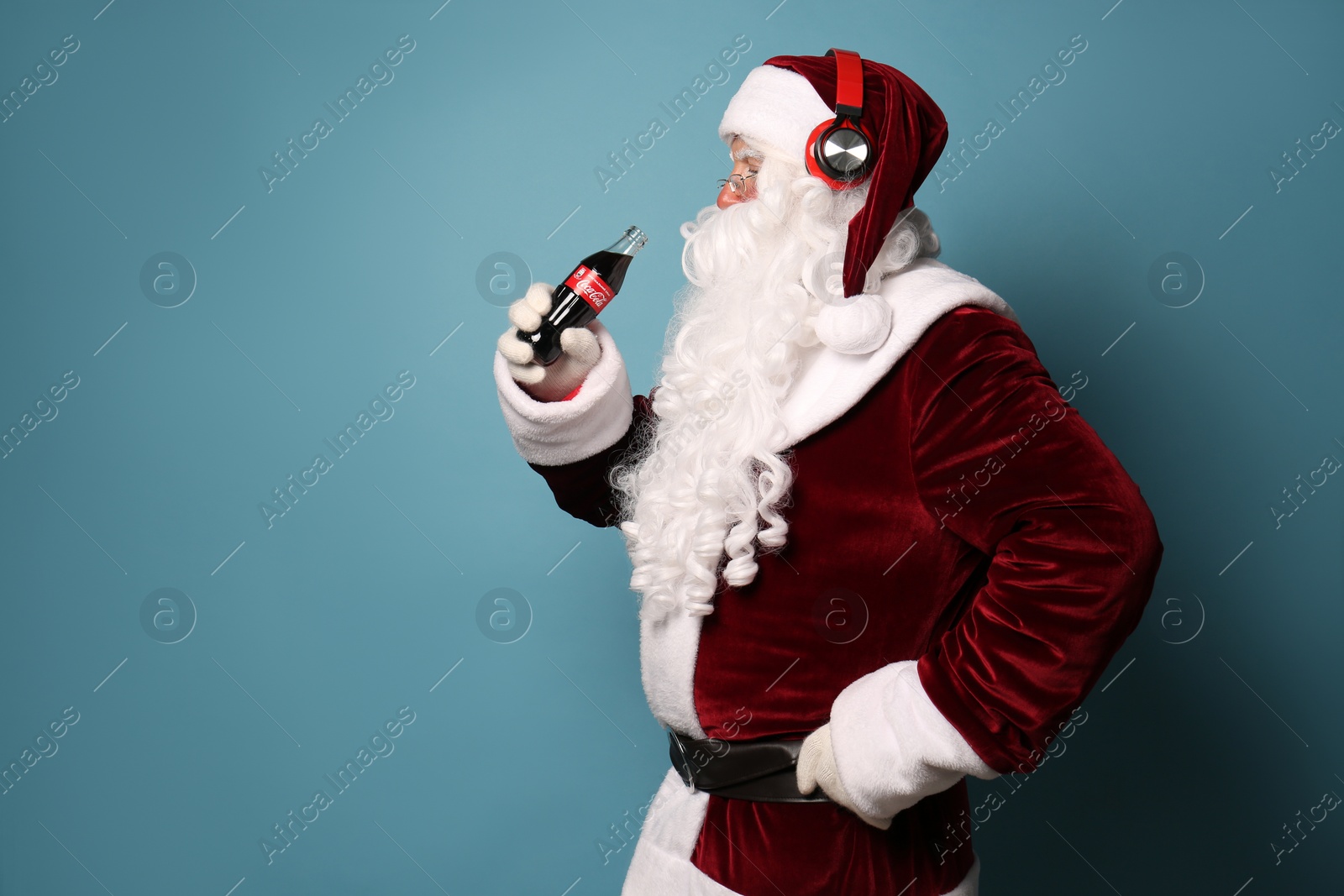 Photo of MYKOLAIV, UKRAINE - JANUARY 18, 2021: Santa Claus holding Coca-Cola bottle and listening to music with headphones on light blue background