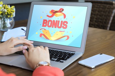 Image of Bonus gaining. Woman using laptop at wooden table indoors, closeup. Illustration of open gift box, word and confetti on device screen