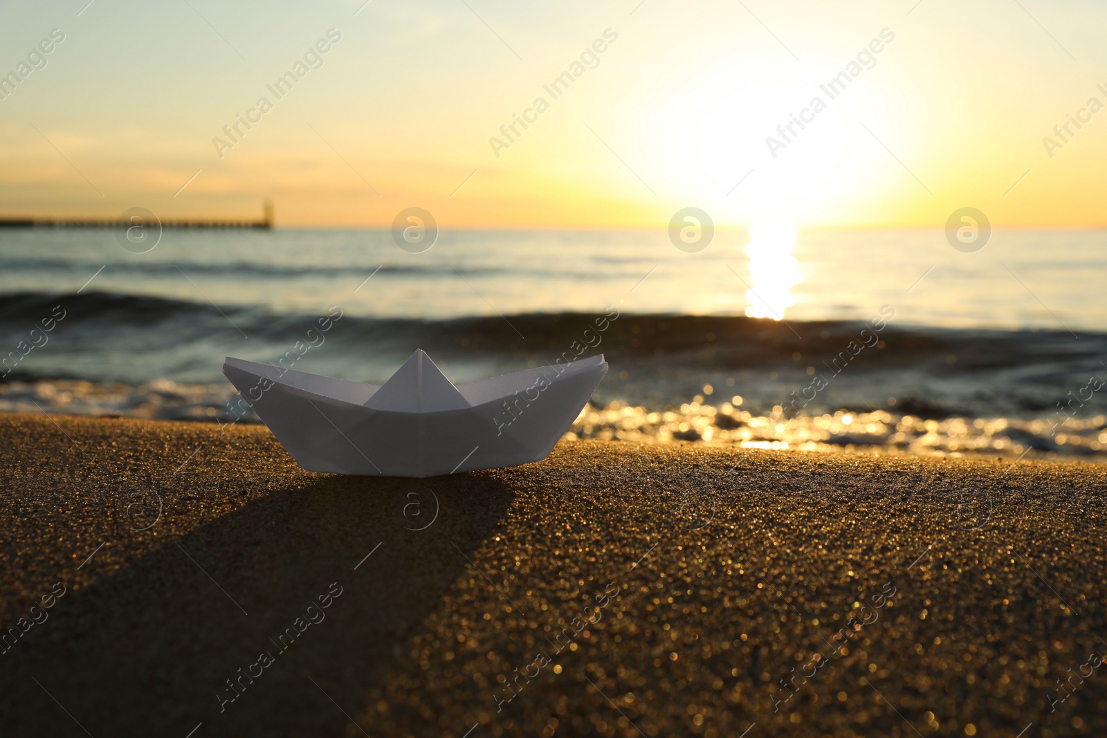 Photo of White paper boat on sand near sea at sunset, space for text