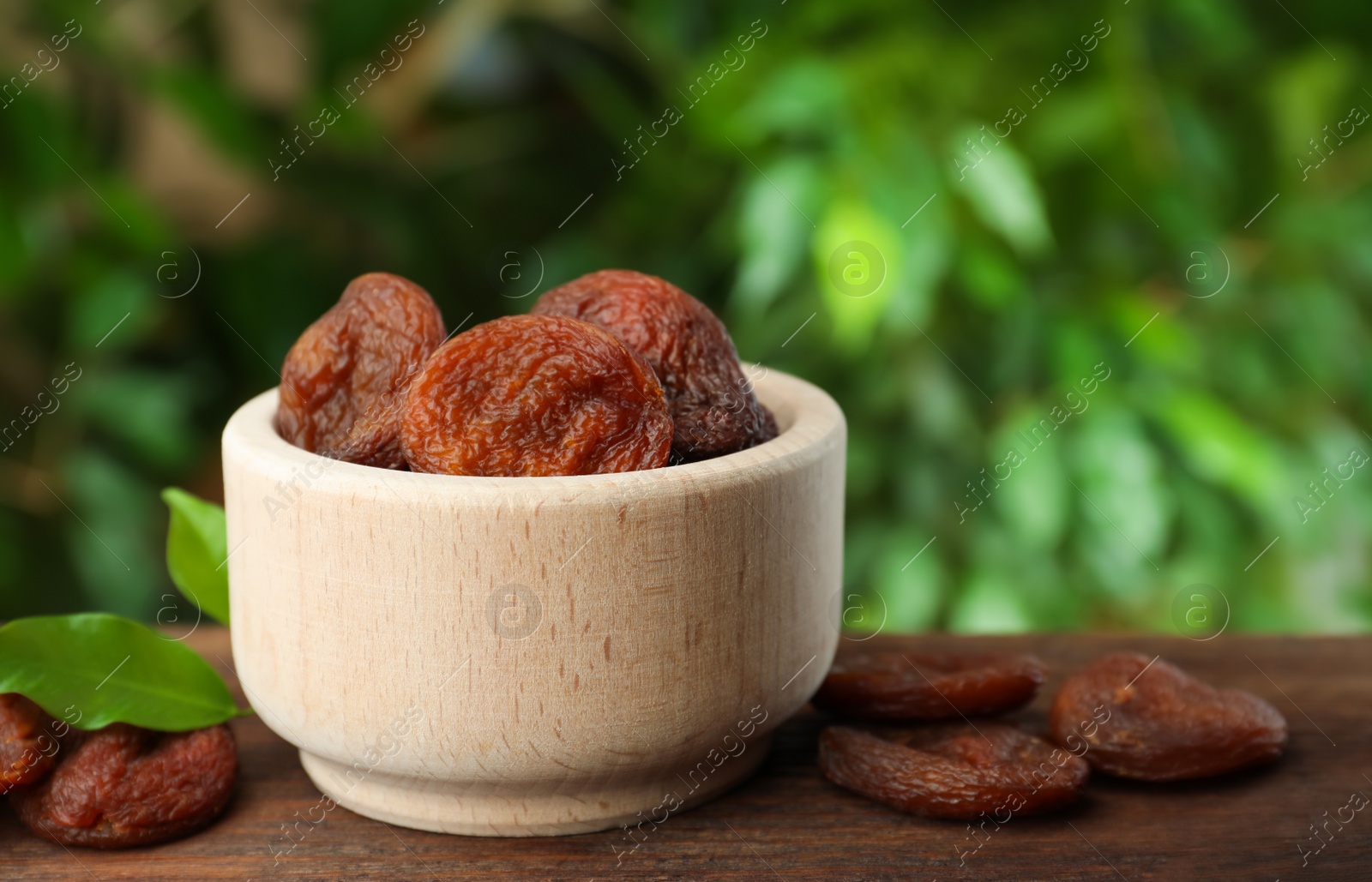 Photo of Bowl of tasty apricots on wooden table against blurred green background, space for text. Dried fruits