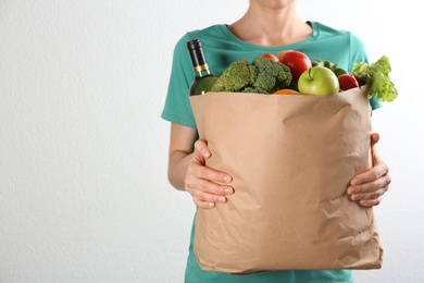 Photo of Woman holding paper bag with different groceries near white wall, closeup view. Space for text