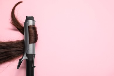 Photo of Curling iron with brown hair lock on pink background, top view. Space for text
