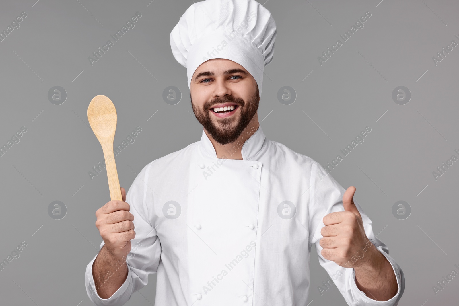 Photo of Happy young chef in uniform holding wooden spoon and showing thumb up on grey background