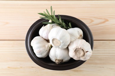 Photo of Fresh garlic bulbs in bowl and rosemary on wooden table, top view