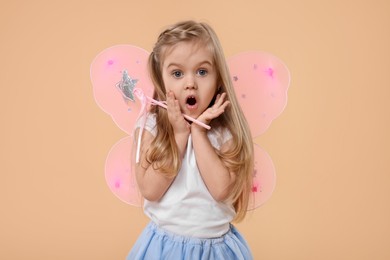 Surprised little girl in fairy costume with pink wings and magic wand on beige background