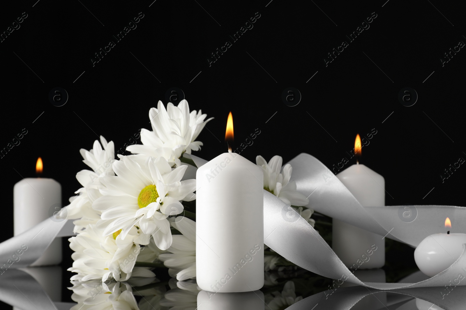 Photo of White chrysanthemum flowers and burning candles on black mirror surface in darkness. Funeral symbols