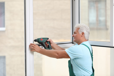 Photo of Mature construction worker repairing plastic window with electric screwdriver indoors