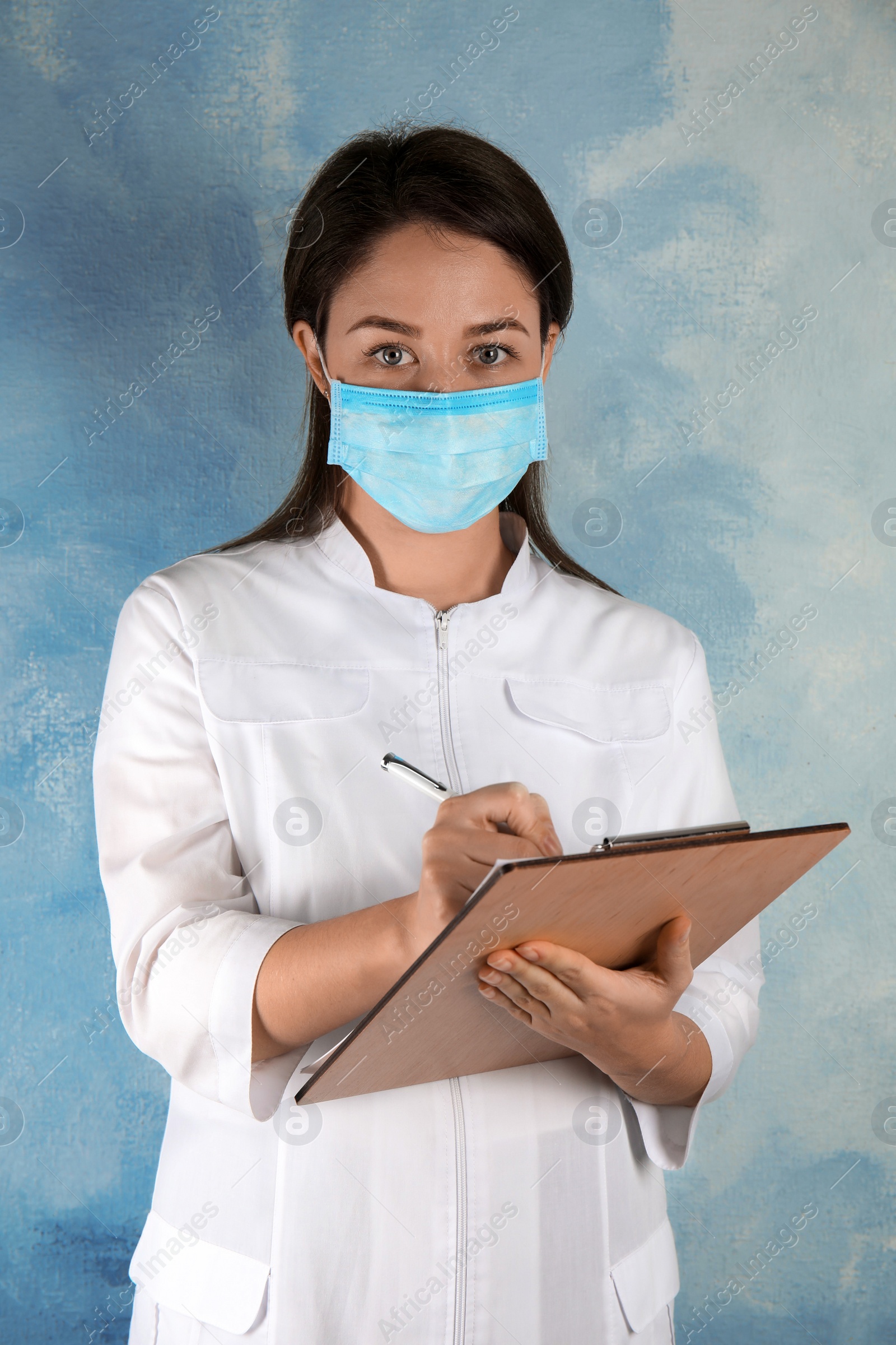 Photo of Doctor with disposable mask on face holding clipboard against light blue background