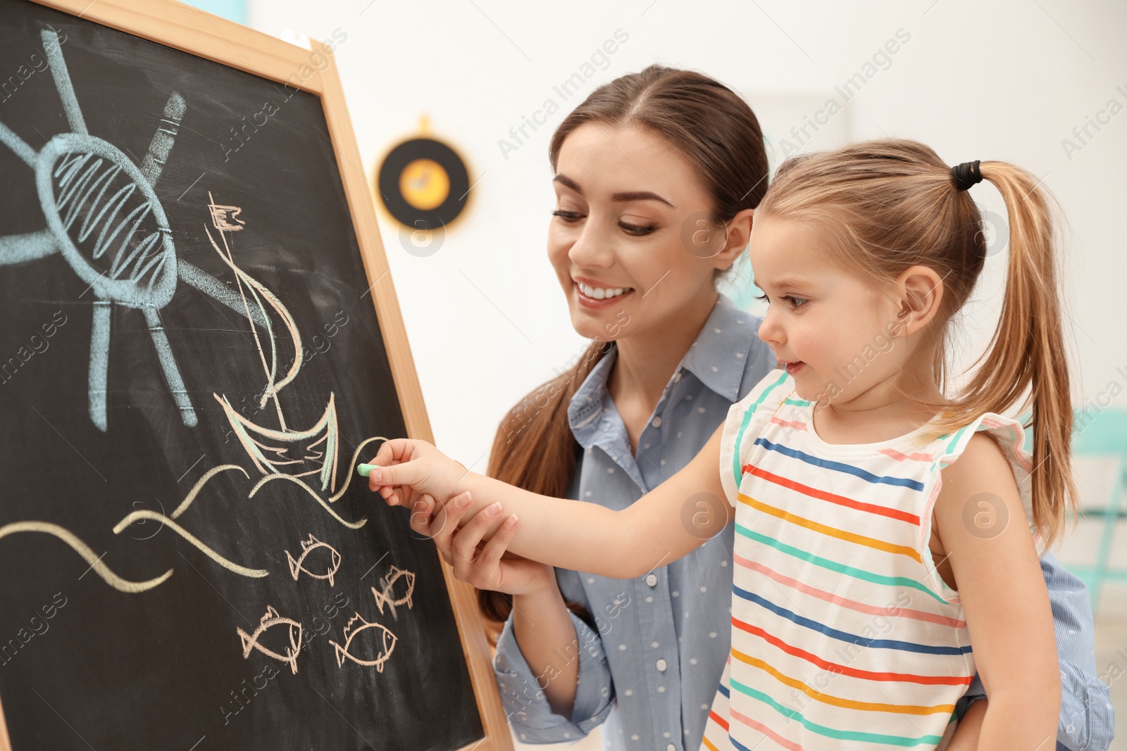 Photo of Kindergarten teacher and little child drawing on chalkboard indoors. Learning and playing