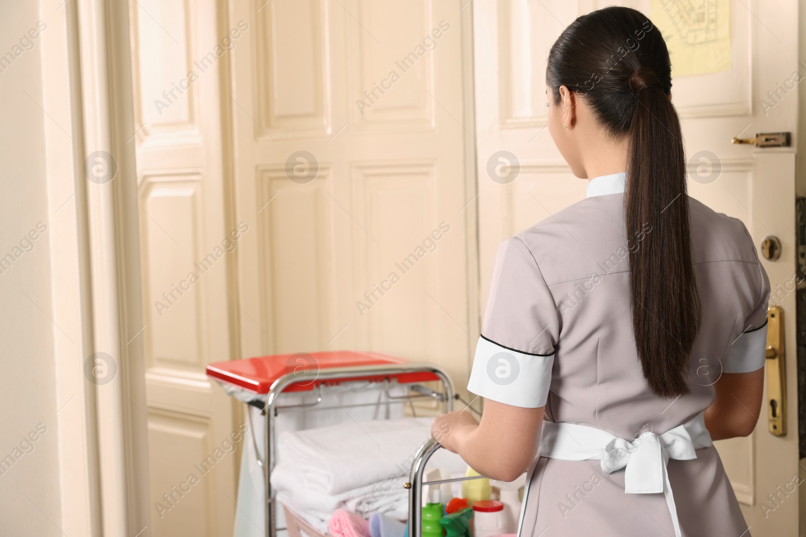 Photo of Young chambermaid with cart and cleaning products in hotel, back view. Space for text