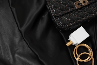 Leather bag, bottle of perfume and golden bracelets on black fabric, flat lay. Space for text