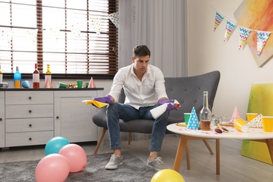 Photo of Tired man with bottle of detergent and rag in messy room after party