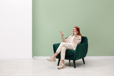 Photo of Happy young woman talking on smartphone while sitting in armchair near light olive wall indoors. Space for text