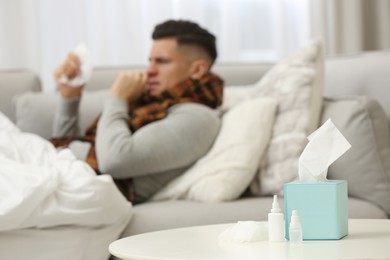 Photo of Ill man at home, focus on nasal spray, drops and box of tissues