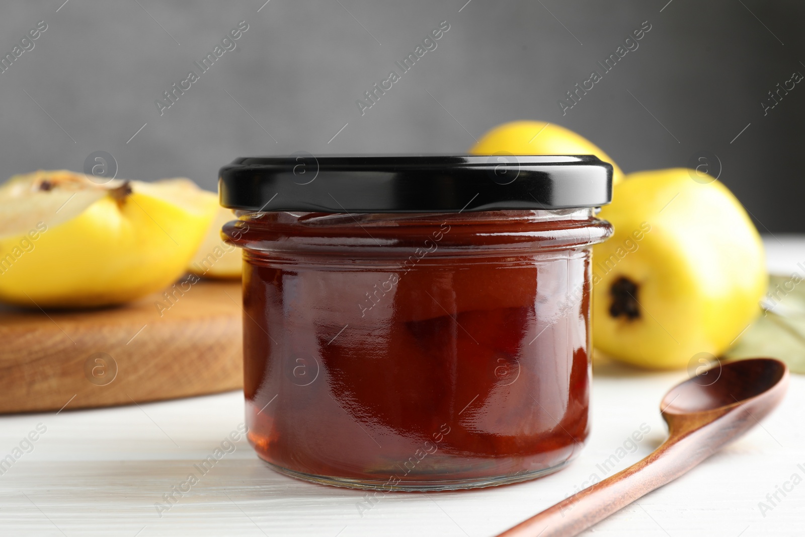 Photo of Tasty homemade quince jam in jar, spoon and fruits on white wooden table, closeup