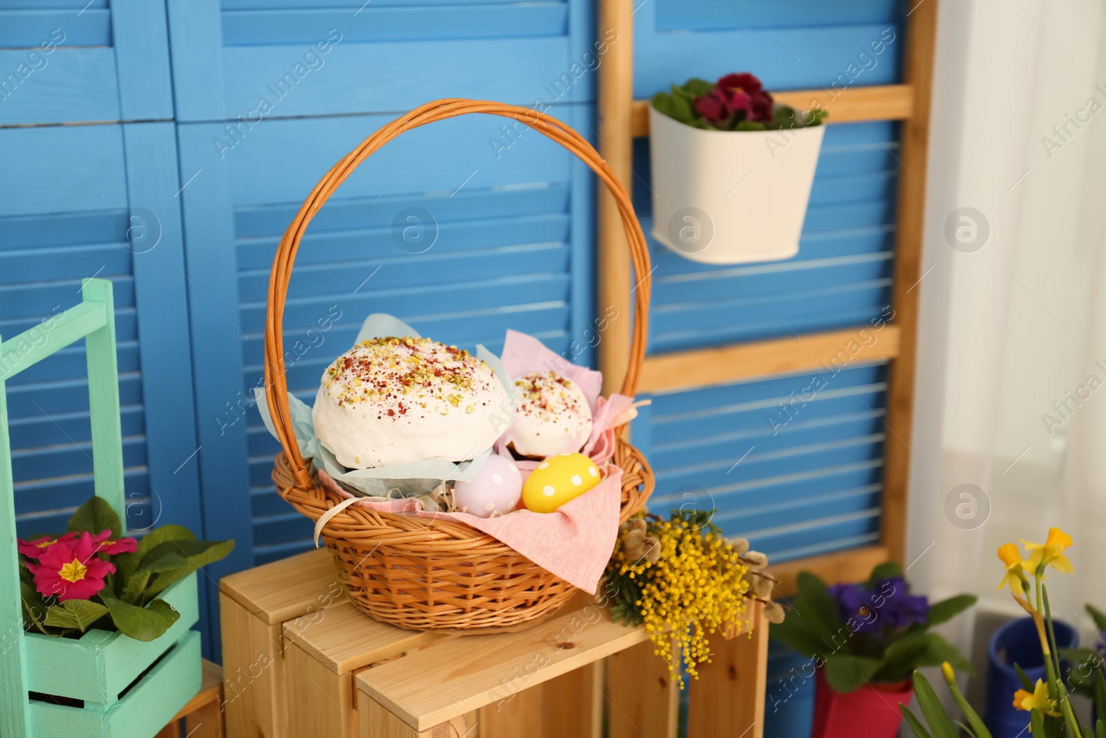 Photo of Basket with delicious Easter cakes, dyed eggs and flowers on wooden crate indoors