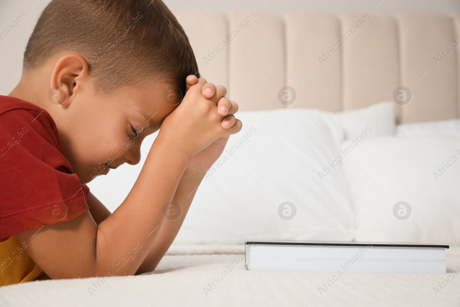 Photo of Cute little boy with hands clasped together saying bedtime prayer over Bible at home. Space for text