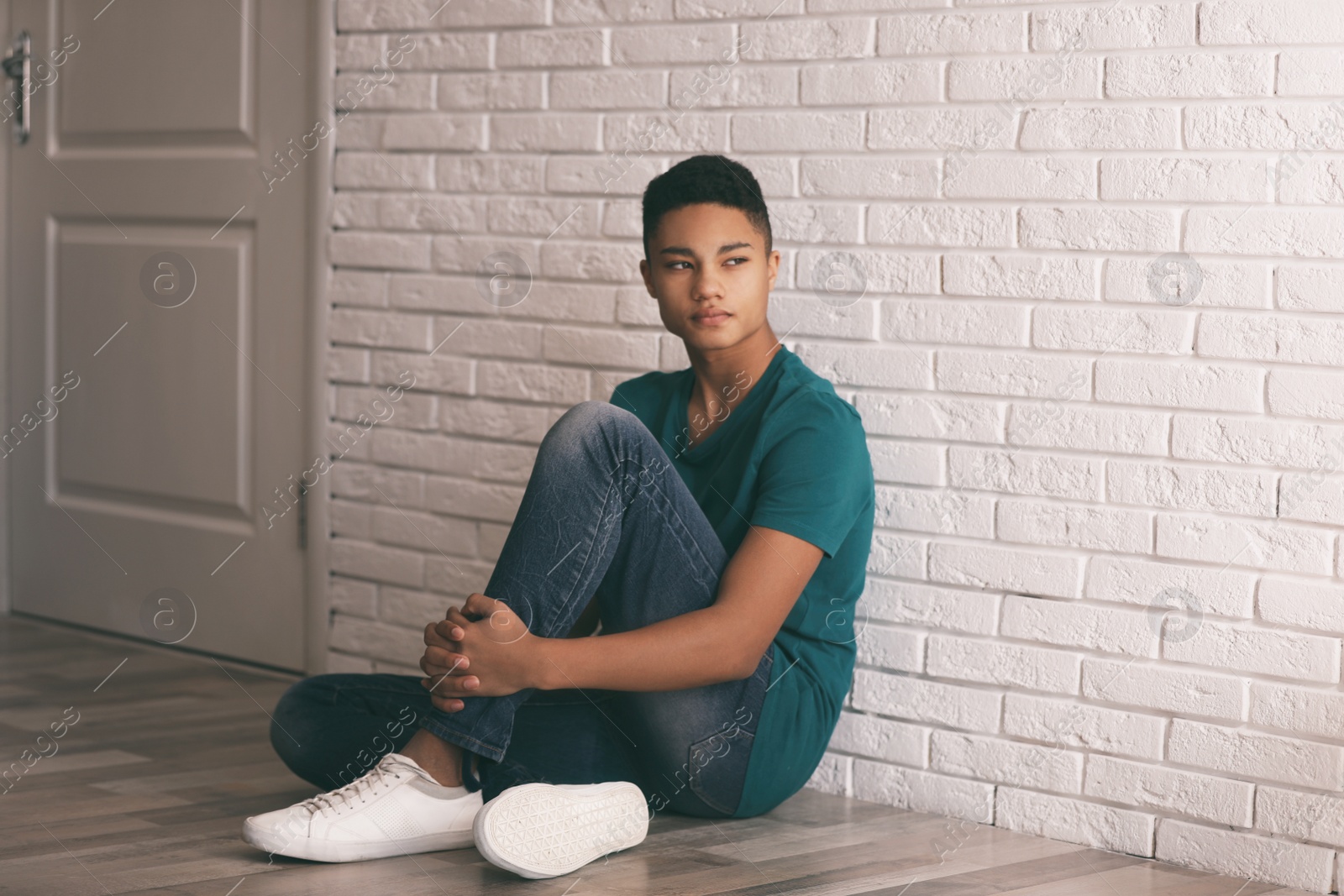 Photo of Upset African-American teenage boy sitting alone on floor near wall