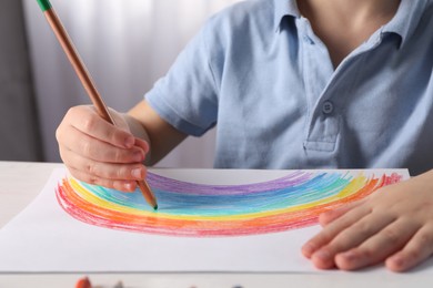Photo of Little boy drawing rainbow with pencil at white table indoors, closeup. Child`s art