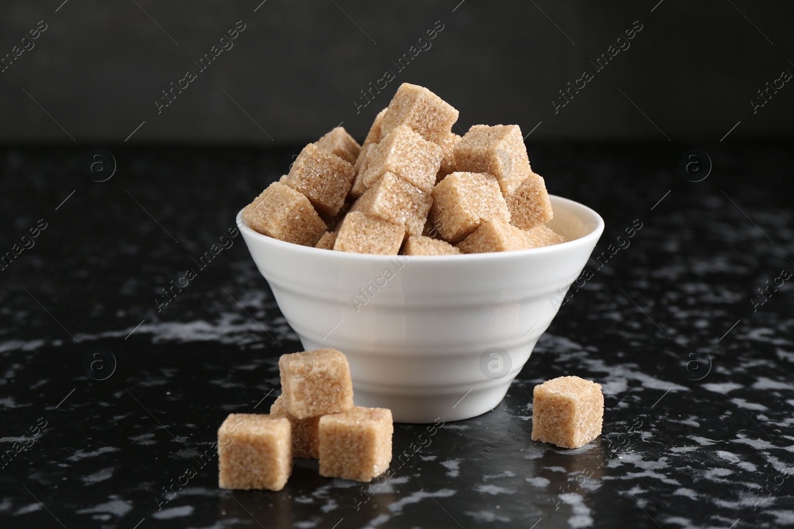 Photo of Brown sugar cubes in bowl on dark textured table, closeup