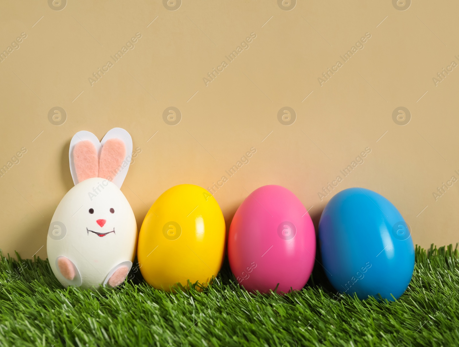 Photo of Bright eggs and white one as Easter bunny on green grass against beige background