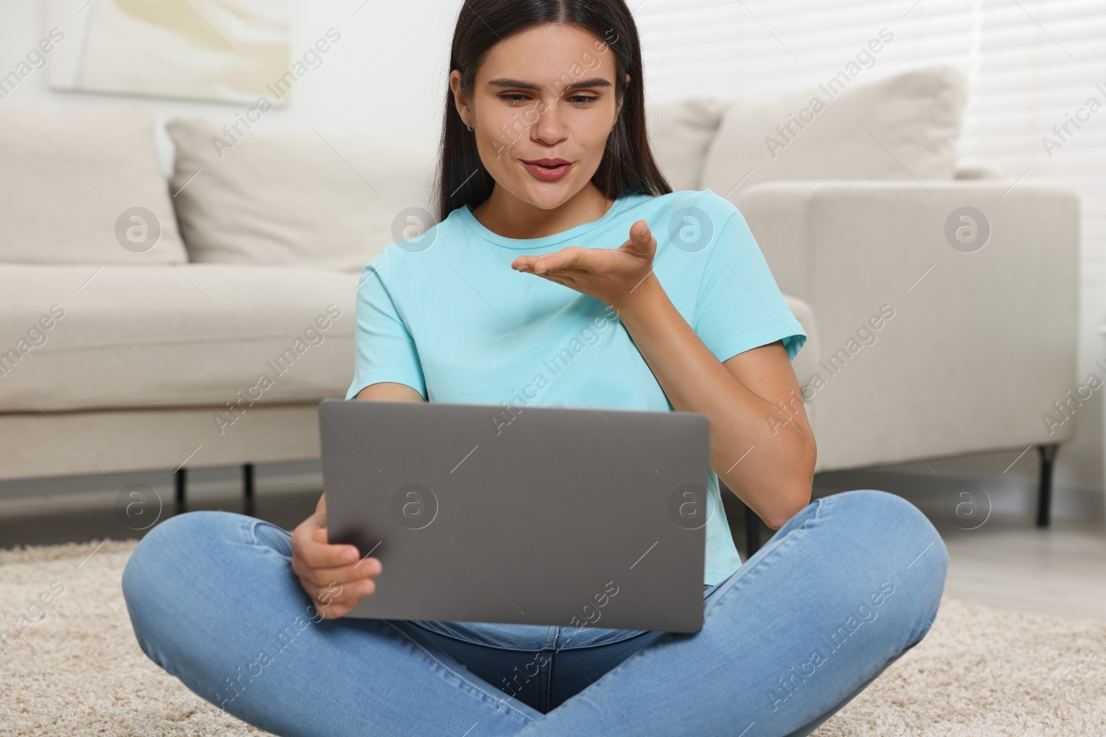 Photo of Young woman having video chat via laptop and blowing kiss on floor at home