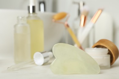 Photo of Jade gua sha tool and toiletries on white countertop in bathroom, closeup