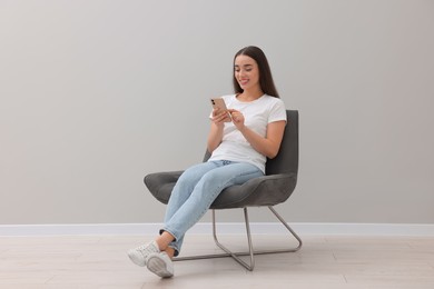 Photo of Beautiful woman using smartphone while sitting in armchair near light grey wall indoors