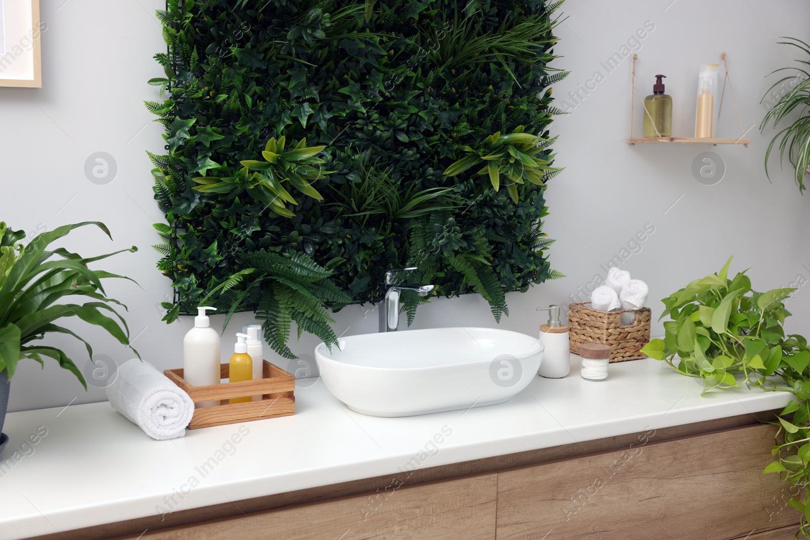 Photo of Green artificial plants, vessel sink and different personal care products in bathroom